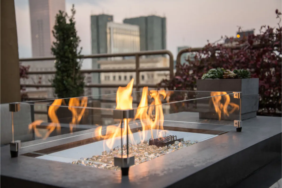 A cozy private outdoor terrace at sunset, featuring a modern gas firepit as the focal point. Succulent planters add a touch of greenery, while the city skyline forms a striking urban backdrop. Comfortable seating is arranged around the firepit, evoking a warm and inviting atmosphere perfect for intimate gatherings or peaceful relaxation.