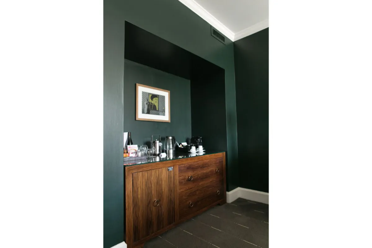 A sophisticated living area with dark green walls, a wooden sideboard, and curated artwork. The space exudes warmth and style, reflecting a local and artistic vibe. It’s set up for both morning coffee rituals and relaxed gatherings.