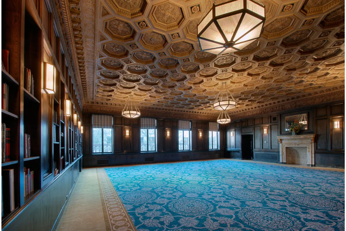 A sophisticated interior space is highlighted by an ornately designed ceiling, a cozy fireplace, grand windows bringing in natural light, and bookshelves filled with carefully curated books. The room exudes an atmosphere of refinement and inspiration, perfect for both productivity and relaxation.