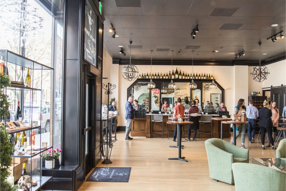 A lively interior featuring a well-appointed bar counter, display shelves with bottles, a comfortable seating area, and guests engaging in relaxed conversation. The ambiance is energetic and welcoming, perfect for social events like wine tastings or mingling during a daily wine hour.