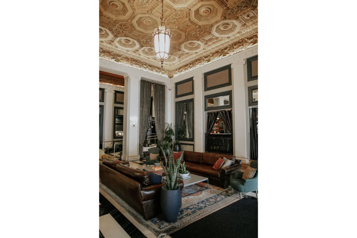 A sophisticated lounge area featuring ornate ceiling details, stylish furnishings, large windows filling the space with daylight, and abundant indoor plants. The arrangement suggests a welcoming setting that blurs the line between indoors and outdoors, promoting relaxation and socializing.