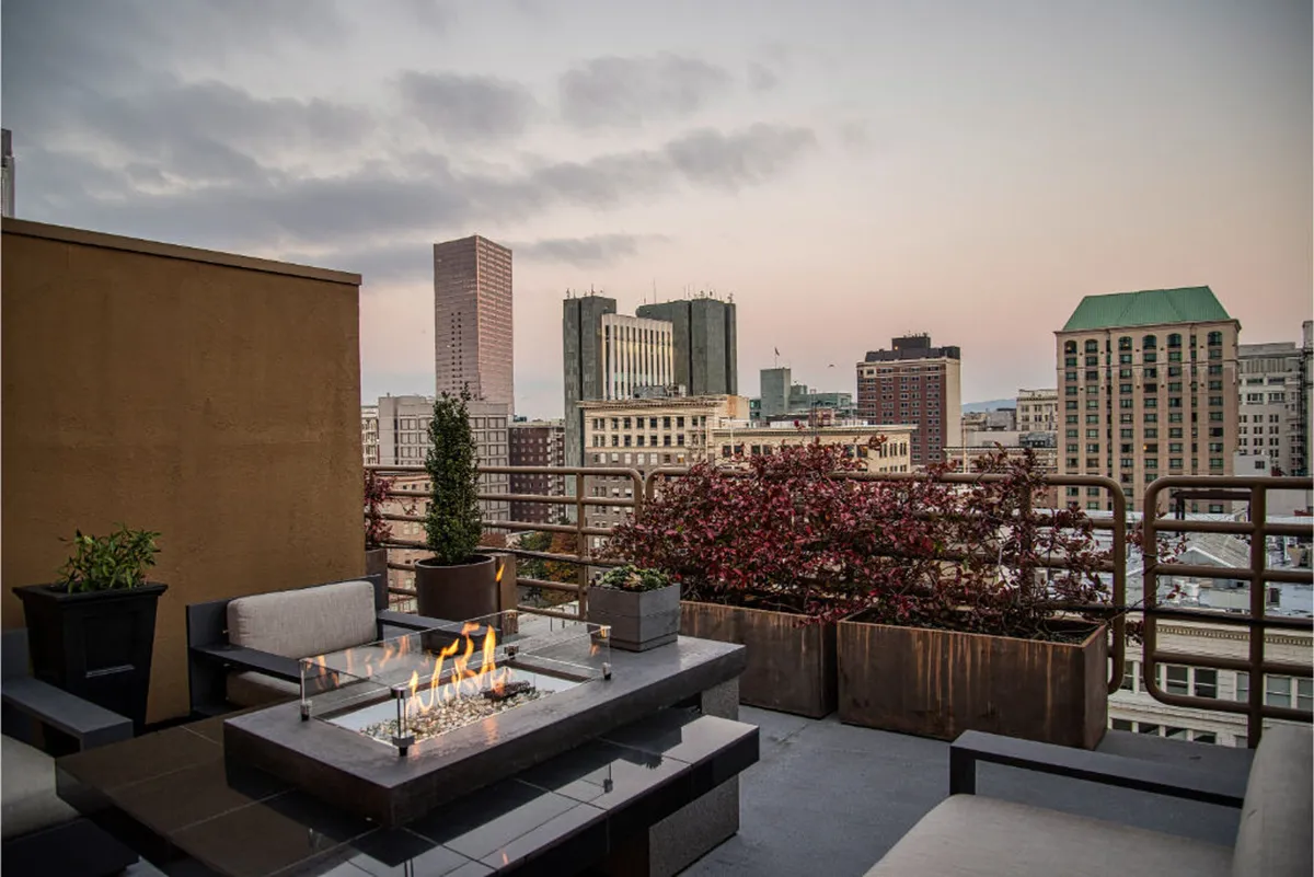 The image showcases the hotel's exterior terrace at dusk, featuring a glowing fire pit surrounded by plush seating, lush planters, and sweeping views of Portland's city skyline. The warm lighting and intimate setting suggest a perfect spot for evening gatherings, relaxation, and enjoying sunset vistas.