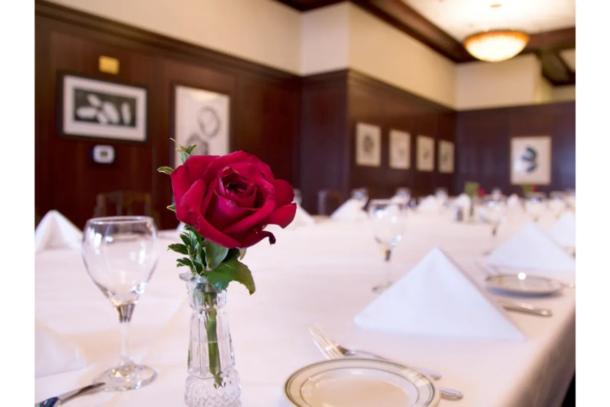 This image features a neatly set dining table with a red rose in a vase, framed artworks on the wall, and an overall inviting and artistic ambiance. The decor is sophisticated yet intimate, with subtle touches that reflect a curated appreciation for art and detail.