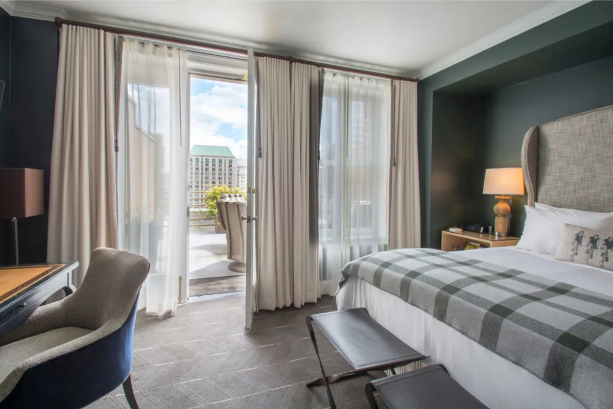 This image features a bedroom with a plush bed adorned with a patterned throw, open balcony doors inviting natural light, a comfortable chair, and a desk with a lamp. The atmosphere is serene and cozy, perfect for both relaxation and productivity.