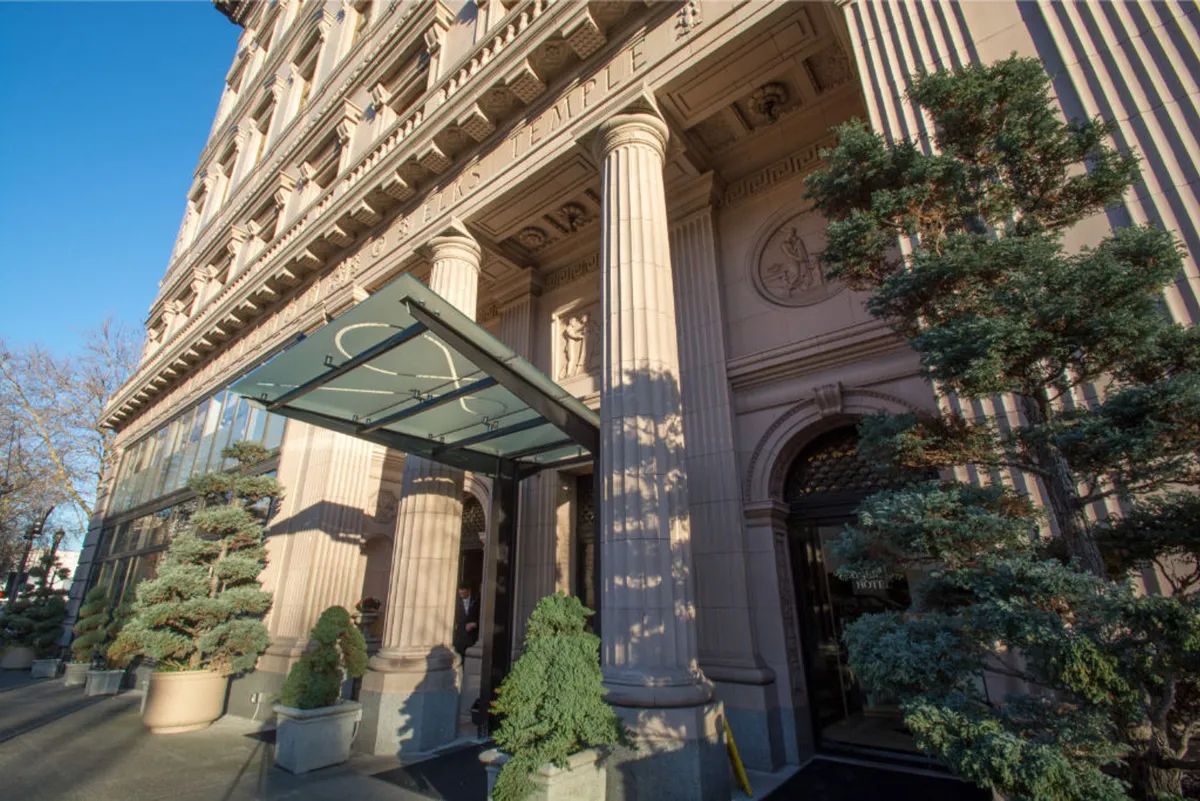 This exterior shot captures the grand entrance of the hotel, featuring ornate columns, a glass awning, and lush, landscaped greenery. The stately facade sets an immediate tone of elegance and exclusivity, inviting guests into a world of luxury and refinement.