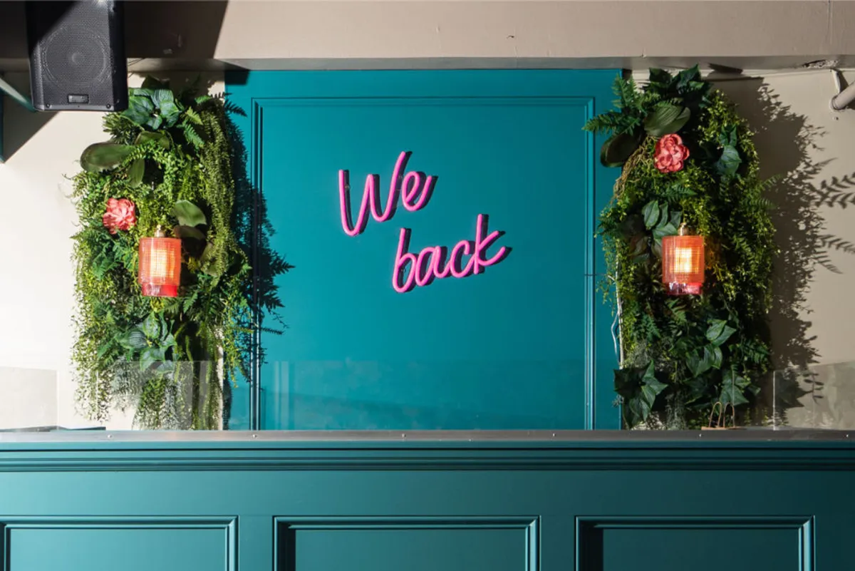 A welcoming, vibrant social space with a neon sign reading 'We back,' lush greenery, and eclectic wall decorations. The scene feels energetic and modern—perfect for gathering before heading out for city adventures.